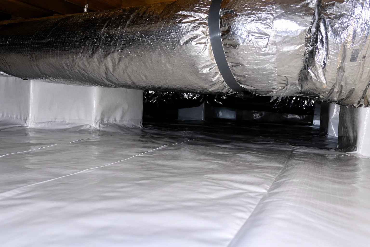 Crawl space with a broad silver insulated duct above a floor covered in white vapor barrier sheeting.