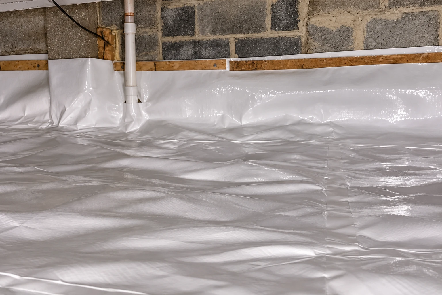 A crawl space floor is covered with a shiny white vapor barrier, with the plastic turned up along the block walls.