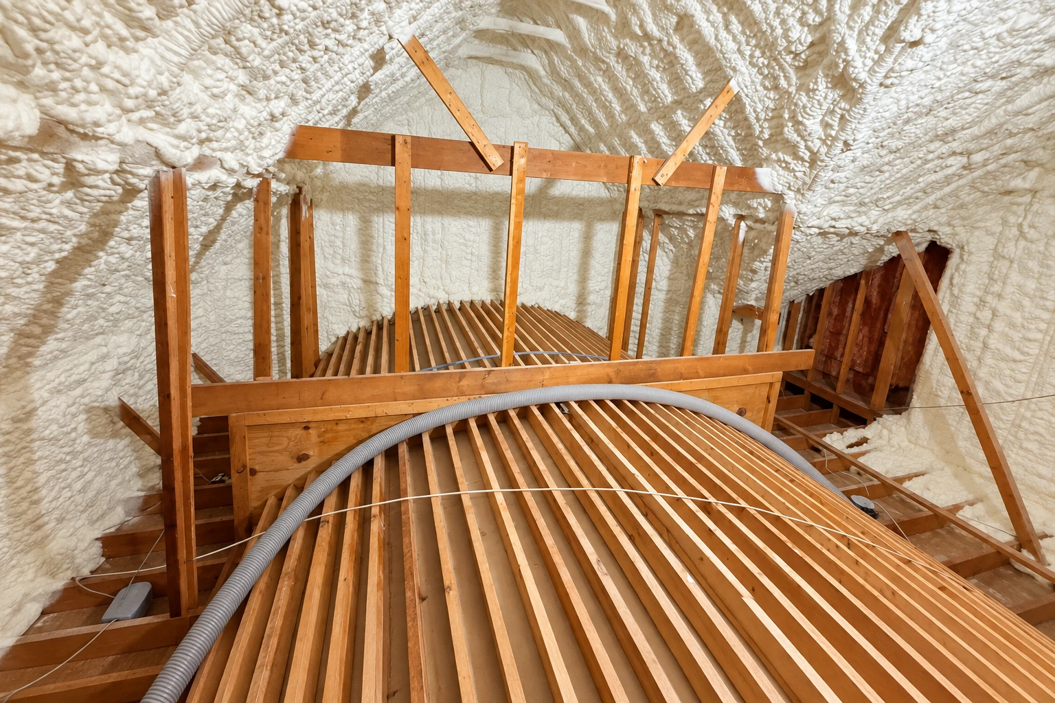 Attic with white spray foam covering the roof and walls above exposed wooden framing and a flexible duct.