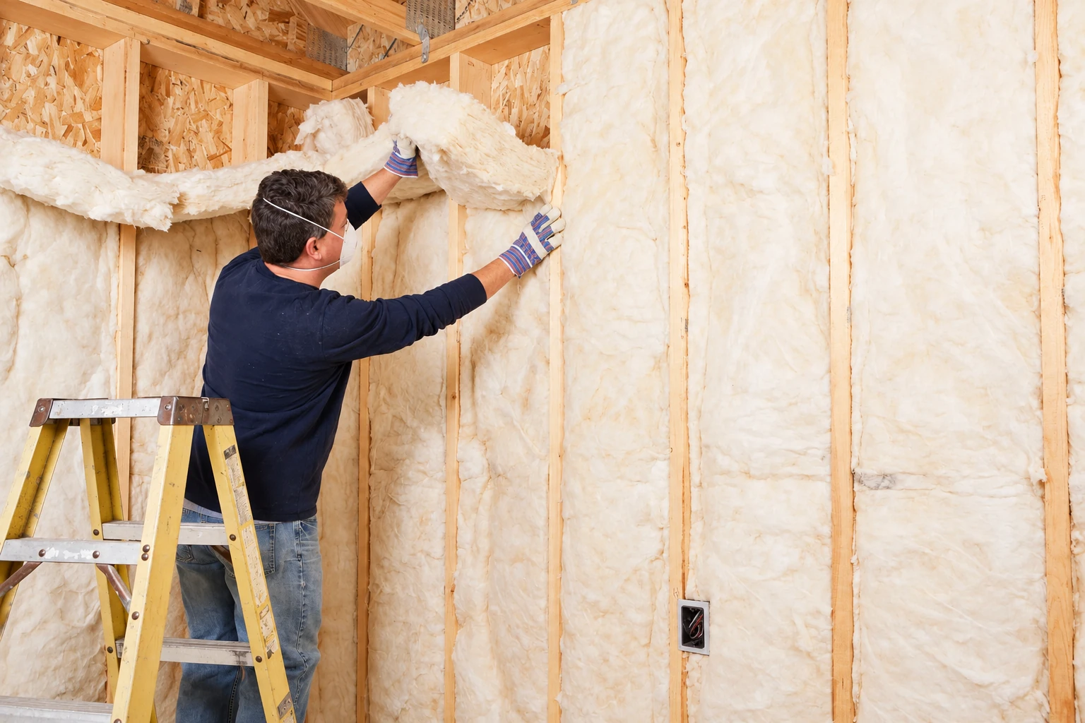 A worker wearing a mask fits fluffy white fiberglass batts between the studs of an unfinished wall.