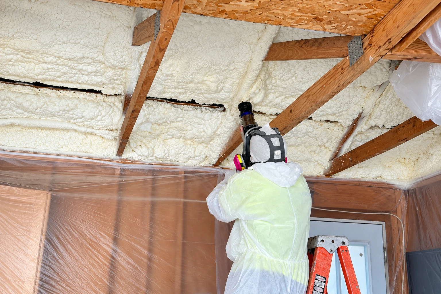 A worker in protective coveralls sprays foam insulation onto exposed roof rafters in an attic.
