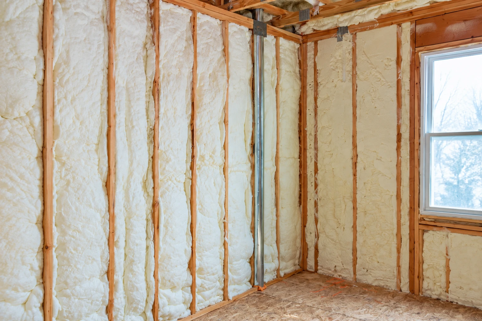Exposed wall cavities are filled with cream-colored foam insulation beside a window.